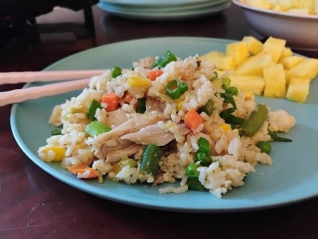 Serving of chicken fried rice on a light blue plate, featuring chunks of chicken, carrots, corn, peas, and green beans, with a side of fresh pineapple cubes and a pair of chopsticks resting on the plate.