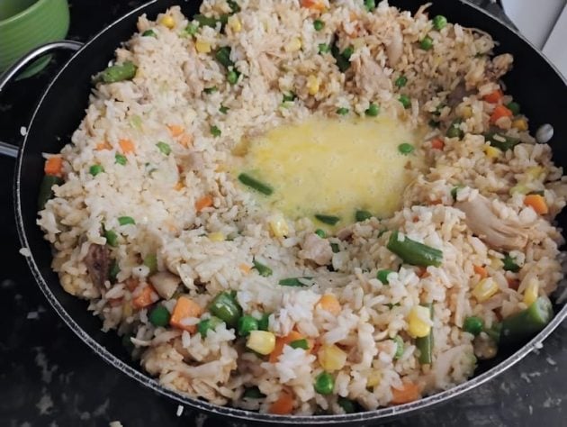 Stir-fry of chicken fried rice with beaten eggs are being cooked, surrounded by rice, carrots, peas, corn, green beans, and chunks of chicken.