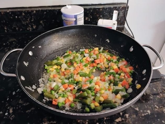 Wok on a stovetop with a colorful mix of sautéed vegetables including carrots, corn, green beans, peas, and onions, cooking together on a black speckled countertop with a container of salt in the background.