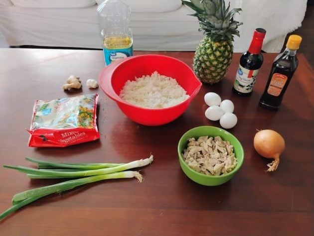 Ingredients for chicken fried rice laid out on a wooden table, including a red bowl of cooked rice, a green bowl of shredded chicken, whole eggs, fresh pineapple, mixed vegetables, soy sauce, teriyaki sauce, sesame oil, cooking oil, green onions, an onion, and pieces of ginger and garlic.