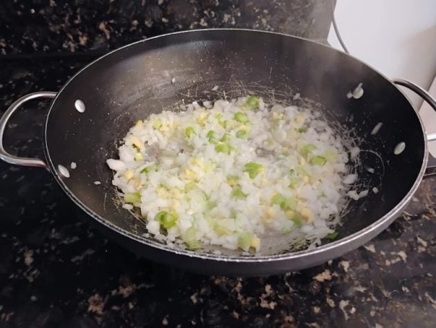 Wok on a stovetop with chopped onions, garlic, and green onions being sautéed in oil.