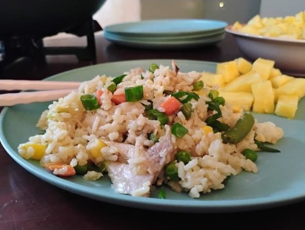 Close-up of a serving of chicken fried rice on a blue plate, featuring diced vegetables, chopped green onions, and tender chicken pieces, with a side of fresh pineapple chunks and a pair of chopsticks resting on the plate.