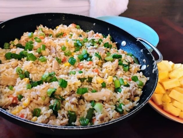 Large skillet filled with homemade chicken fried rice, featuring chunks of chicken, diced vegetables, and garnished with chopped green onions, served alongside a plate of fresh pineapple cubes and a light blue plate in the background.